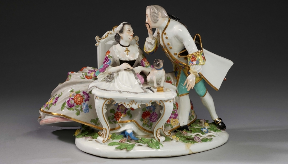 grupa figurek porcelanowych przedstawia mężczyznę i kobietę w strojach z epoki, na stoliku za którym siedzi kobieta jest mały piesek
