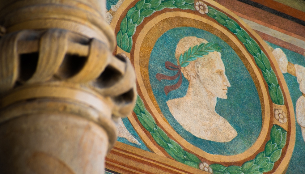 fragment kolumny na krużgankach wawelskich, ozdobionej elementem rzeźbiarskim w kształcie plecionki, w tle barwne malowidło przedstawiające portret mężczyzny w wieńcu laurowym